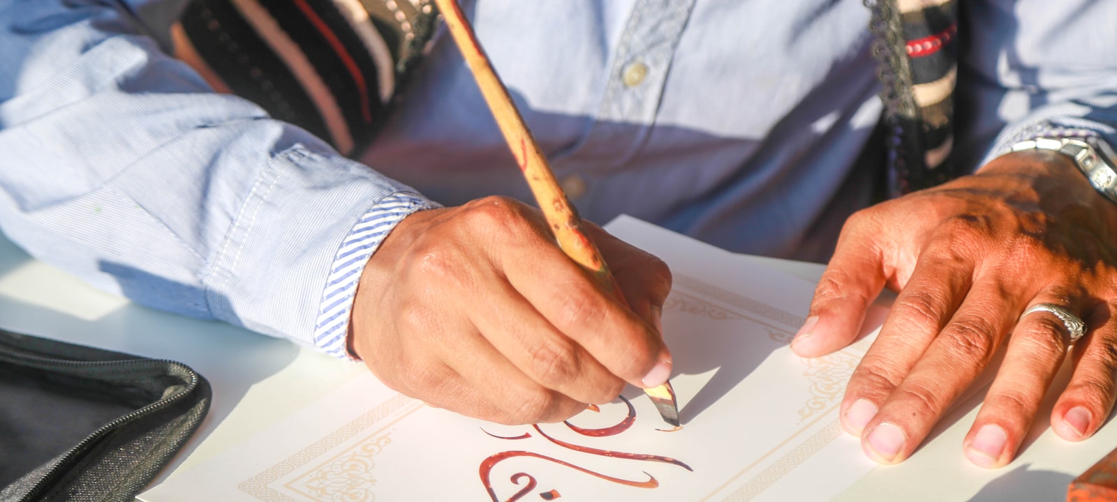 A calligrapher writing Arabic script with a reed pen on ornamented paper