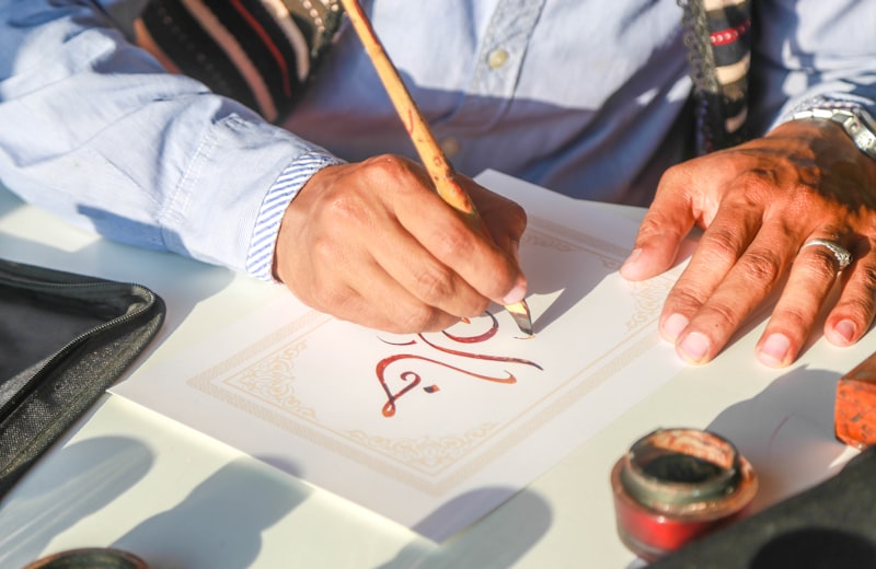 A calligrapher writing Arabic script with a reed pen on ornamented paper